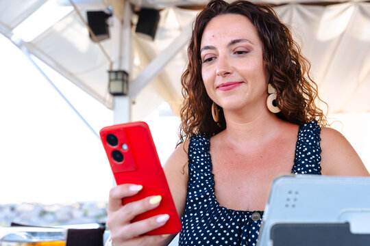 Smiling woman using smartphone with red case while working on tablet at outdoor cafe, modern lifestyle with digital technology, communication, leisure, business, internet connection, summer casual