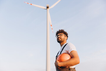 Technician holding helmet looking up at wind turbine in sustainable energy plant
