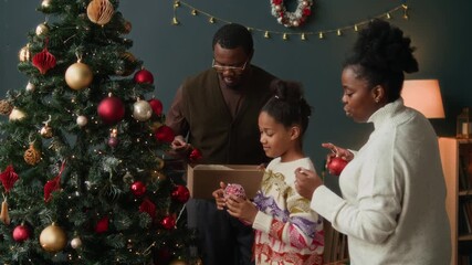 Focused African American girl child helping parents decorating Christmas tree hanging red baubles on branches of artificial fir tree while enjoying winter holidays with family at home - Powered by Adobe