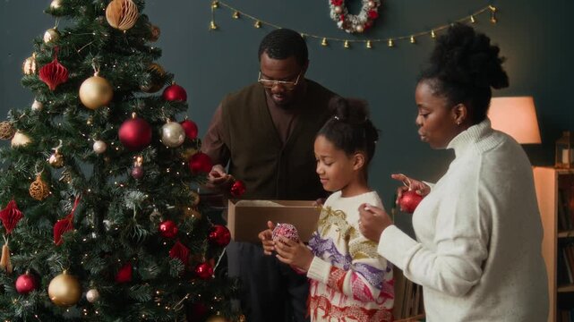Focused African American girl child helping parents decorating Christmas tree hanging red baubles on branches of artificial fir tree while enjoying winter holidays with family at home