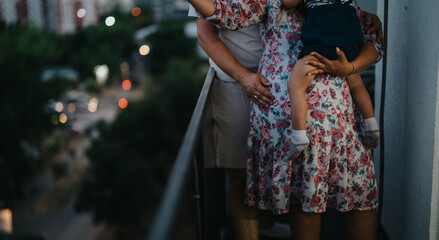 A family stands on a balcony during twilight, embracing a small child while city lights glow softly in the background. A warm moment of togetherness and care outside the home. © qunica.com