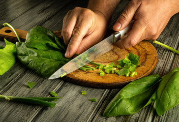 Hands skillfully chop fresh spinach on a wooden board, preparing ingredients for a healthy meal in a bright kitchen. The focus is on the knife and vibrant greens