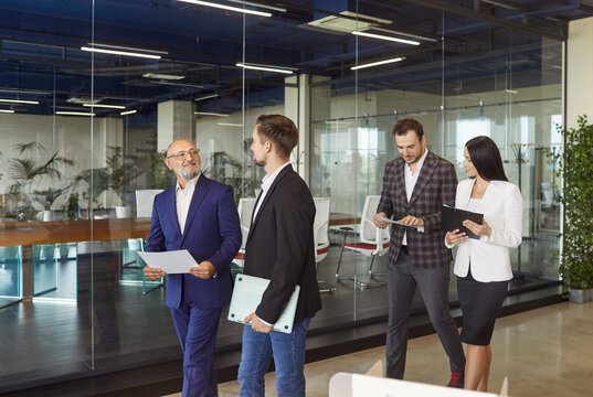 Business people team walking together in modern glass office corridor to discuss corporate meeting, financial data in paper documents. Group of busy professional employees brainstorming