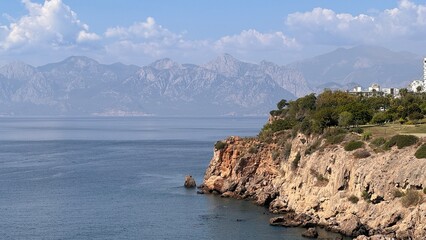Panoramic view of the steep orange-rock Mediterranean sea cliff in Antalya, with lush green parkland above and the majestic Taurus Mountains on the distant horizon