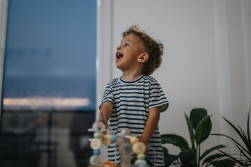 A cheerful toddler in a striped shirt enjoys imaginative play with a pastel bead toy, indoors near green plants and a large window, capturing warmth, joy, and family-friendly moments.