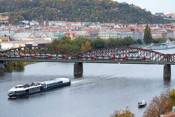 Scenic view of a river with a boat cruising beneath a historic bridge, surrounded by colorful autumn foliage and charming city architecture in the background, showcasing urban beauty