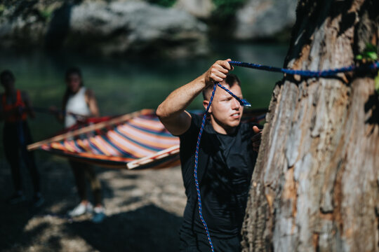 A man prepares a hammock in nature beside a river with friends assisting nearby. This outdoor activity showcases teamwork and enjoyment of the natural environment, promoting relaxation and adventure. - Powered by Adobe