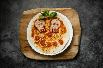 Close-up of Halloween-themed pizza with skull-shaped eyes and tomato mouth on white plate over wooden board, top view food photography for spooky presentation