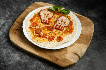 Close-up of Halloween-themed pizza with skull-shaped eyes and tomato mouth on white plate over wooden board, top view food photography for spooky presentation
