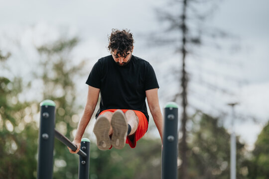 A man in a black shirt and red shorts trains on outdoor parallel bars, focusing on core strength and balance. The park setting provides a blurred, natural backdrop for this fitness moment.