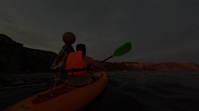 Kayaking ocean night, person paddles past rocky island in dark sea towards distant city lights