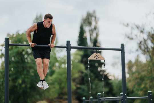 A fit man in a black sleeveless top and shorts grips parallel bars at an outdoor gym, concentrating on his pull-up as green trees form a calm park backdrop.