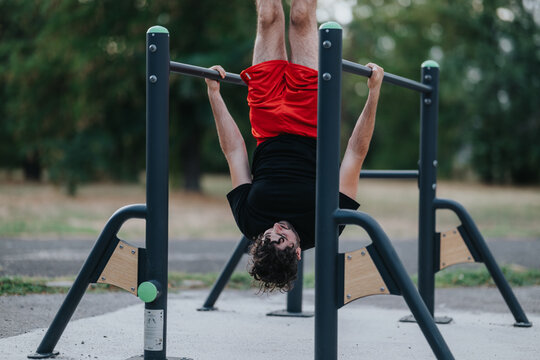 A man in red shorts and black shirt performs an inverted hang on an outdoor fitness bar in a park, showcasing strength, balance, and an active outdoor workout. - Powered by Adobe