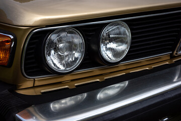 Close-up of the headlights of a beige European car from the 1980s. Front corner of the car caught in daylight.