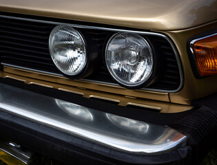 Close-up of the headlights of a beige European car from the 1980s. Front corner of the car caught in daylight.