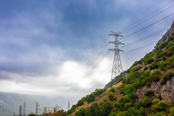 High voltage power lines on a hillside under a cloudy sky. The landscape features green vegetation...