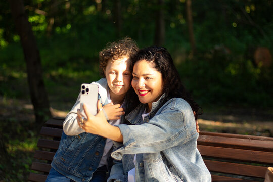 Young mother making selfie or video call with her son sitting on bench in park. Mom and boy calling grandparents. Concept of: connection, distance, technology