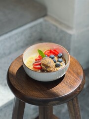 Yogurt granola fruit bowl with peanut butter and berries on wooden stool, side view, sunlight