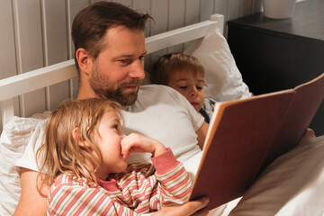 Happy mature 40 year old father reading book to his curious children lying in a bed before the...