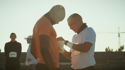 Two male runners share a laugh while one helps the other attach his race bib. They are preparing for a race event, showcasing camaraderie and teamwork before the competition begins.