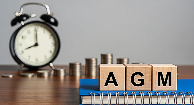 Agm wooden blocks sit atop a blue notebook with coins and a clock in the background representing annual general meeting time