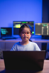 Young girl with glasses working on a laptop in a tech-savvy workspace with multiple computer...