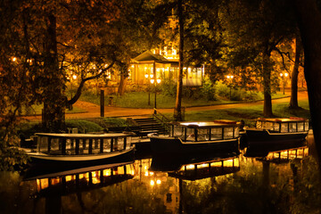 Three pleasure boats are moored in a serene lake at Bastejkalns Park. The scene is illuminated by...