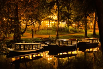 Selbstklebende Fototapeten Fallen Three pleasure boats are moored in a serene lake at Bastejkalns Park. The scene is illuminated by warm lights from nearby trees and a building.  © Kyrylo