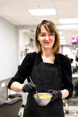 A smiling young woman hairdresser in black rubber protective gloves stirring hair dye with a brush at a beauty salon. Professional hair care, coloring, and coloristics. Selective focus.