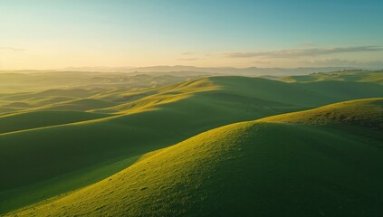Obraz premium Rolling grassy hills glowing under soft golden light with distant mountains under pale blue sky