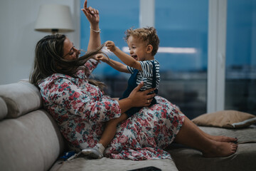 A joyful mother in a floral dress plays with her happy toddler on a sofa in a cozy living room. They share laughter, movement, and close bonding in a warm home scene.