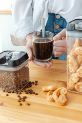 Woman hands, holding a transparent glass with coffee, coffee beans in transparent container. Kitchen table.