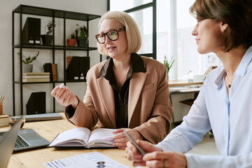 Caucasian senior woman discussing business strategy with middle aged Caucasian woman in modern office setting, both sitting at desk with open notebook and digital tablet visible