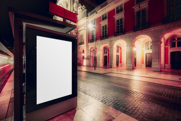 Blank vertical advertising billboard at night on empty city street with illuminated historic architecture, cobblestone pavement, and warm urban lights creating cinematic atmosphere; copy space area