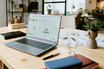 Open laptop displaying business analytics charts on wooden desk in modern office, surrounded by documents, notebooks, glass of water, potted plants, and office supplies, no people visible