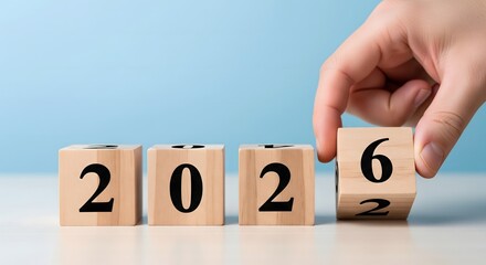 A symbolic image showing the transition from the year 2025 to 2026, represented by wooden cubes with black numbers and a hand flipping the last cube to reveal 