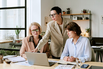 Three middle aged Caucasian women collaborating at desk, one standing and pointing at laptop screen while two others sitting and listening attentively during business meeting
