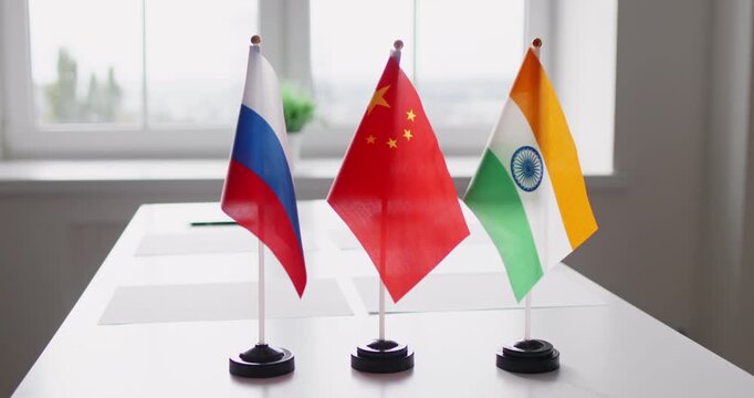 Diplomacy flags indicate negotiation and trilateral cooperation. Three national flags stand on a clean office table, suggesting organized talks. Concept: strategic trilateral partnership.