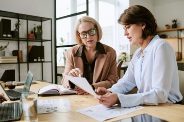 Two middle aged Caucasian women discussing documents at desk, collaborating on business project...
