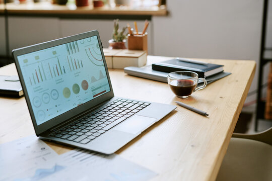 Open laptop displaying business analytics dashboard on wooden desk with documents, pen, cup of tea and notebooks, suggesting workplace setup for remote work or data analysis