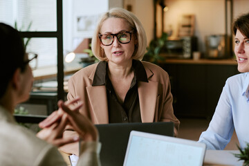 Caucasian middle aged woman wearing glasses engaging in business meeting with two colleagues in modern office setting, discussing project while sitting at table with laptops visible