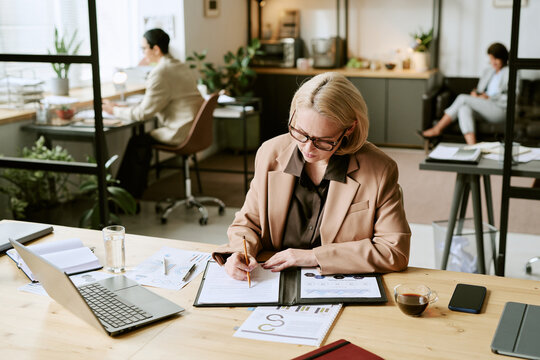 Caucasian middle aged woman working at desk, reviewing documents and writing notes, using laptop and surrounded by office supplies, business charts and coffee, modern office setting - Powered by Adobe