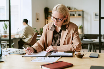 Middle aged Caucasian woman working at desk reviewing documents and making notes with pen, wearing eyeglasses, sitting in modern office with digital tablet and coffee nearby