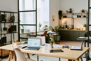 Modern office workspace featuring wooden desk with open laptop displaying charts, digital tablet, smartphone, documents, potted plants, shelving units, kitchen area in background