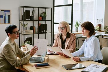 Three middle aged Caucasian women sitting at table discussing business documents in modern office, holding papers and using laptop, collaborating on project together