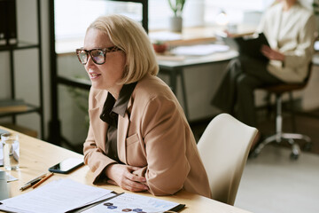 Caucasian middle aged woman sitting at desk smiling during business meeting, wearing eyeglasses,...