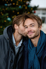 A heartwarming scene of a man smiling as he receives a kiss from another, highlighting the warmth of affection amidst a cold winter backdrop with festive decorations.