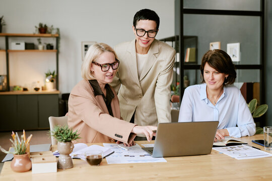 Three middle aged Caucasian women collaborating around laptop in modern office, one standing and smiling while two sitting and discussing project, documents and coffee on desk