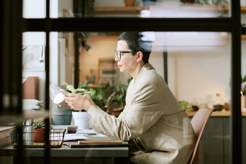 Caucasian middle aged woman sitting at desk working with documents and holding smartphone,...