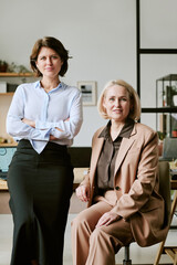 Caucasian young adult woman standing with arms crossed next to seated Caucasian middle aged woman...